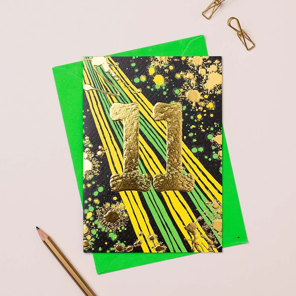 A colorful birthday card with gold foiling and a number 11, designed for an 11-year-old boy, displayed on a table with a green envelope and a pencil.