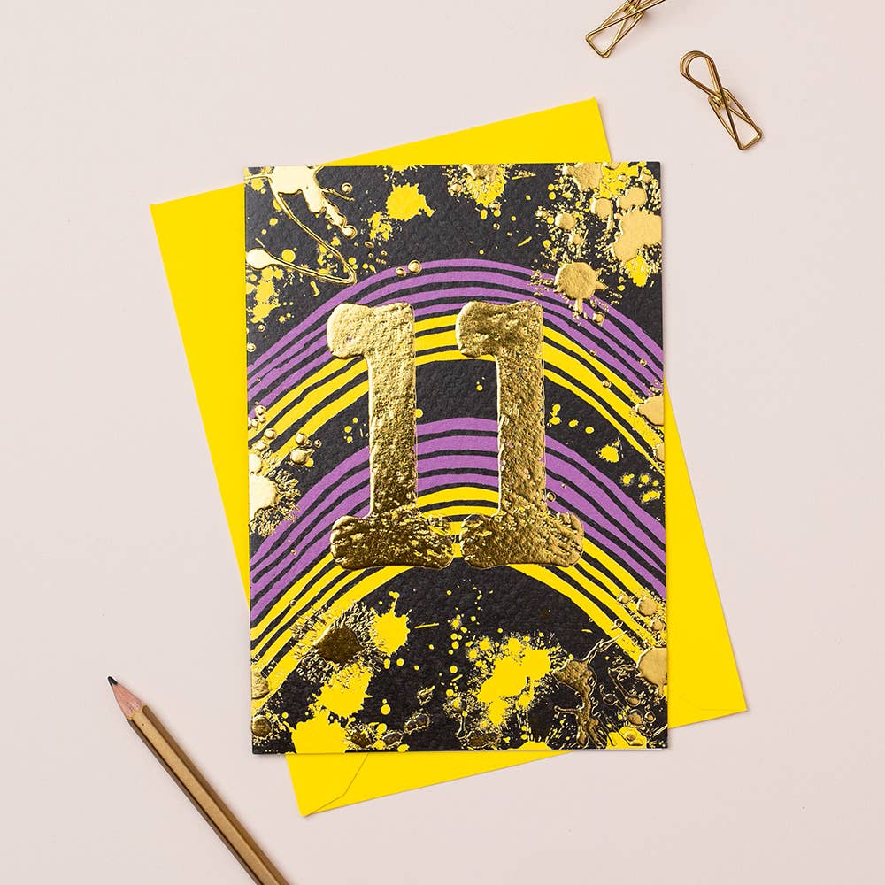 A vibrant birthday card with a gold foil splash and the number 11, placed on a yellow envelope, with a pencil and paper clips on the side.
