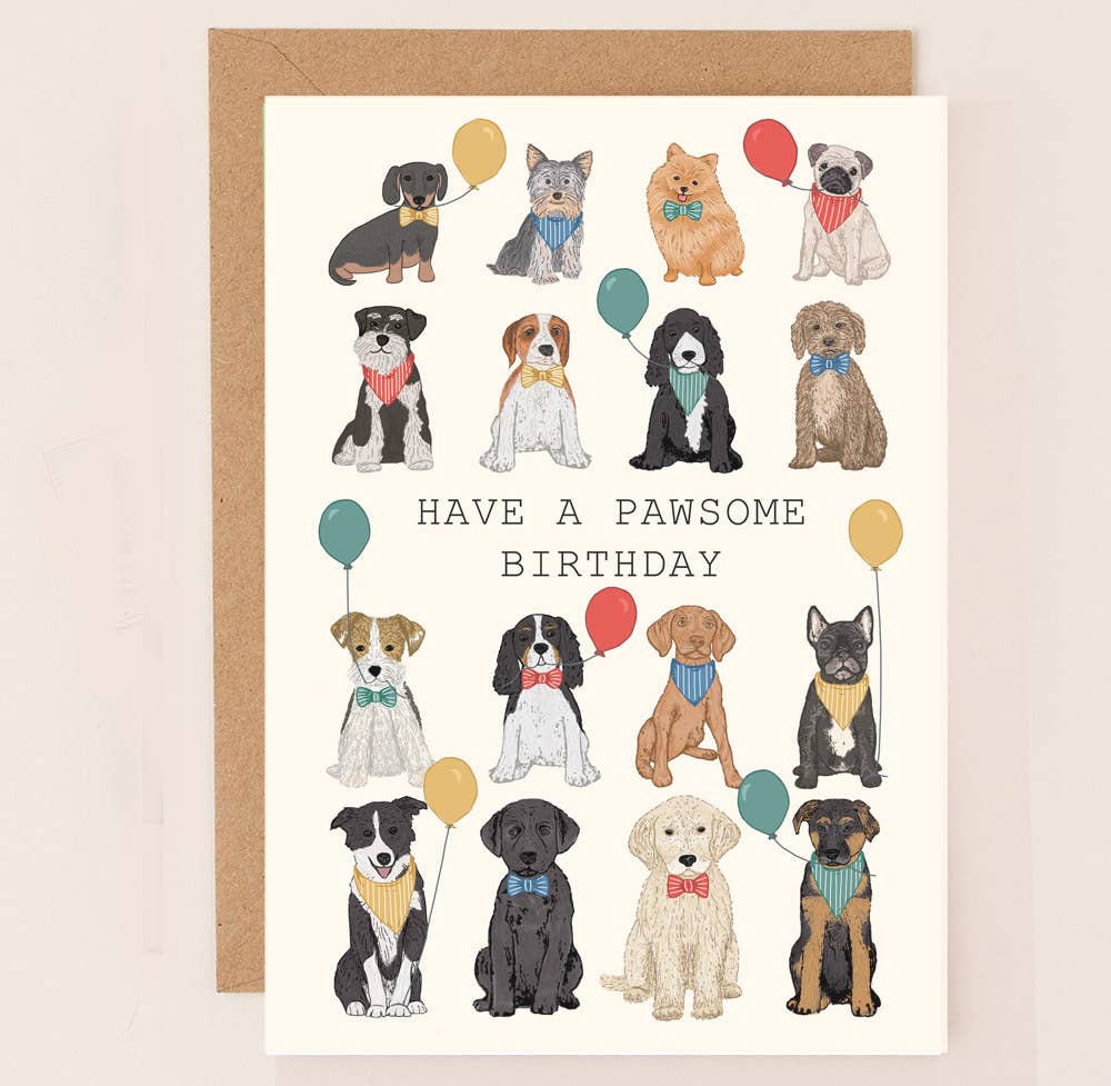 A birthday card with various dog illustrations, each wearing a bow tie and holding a balloon, with the text 'HAVE A PAWSOME BIRTHDAY' at the center. The card is accompanied by a brown kraft envelope.