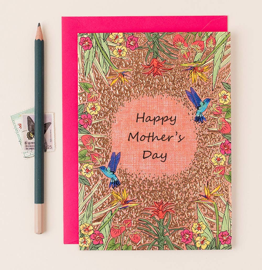 A Mother's Day card with a gold foil border and tropical flower design, featuring hummingbirds and the text 'Happy Mother's Day'. Accompanied by a red envelope and a pencil, all placed on a white surface.