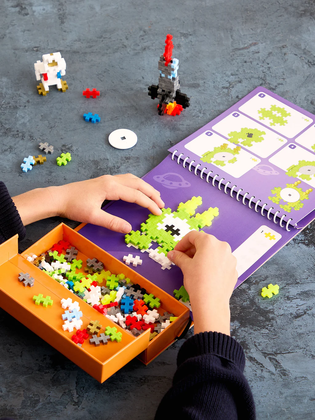 Children playing with colorful building blocks and a purple activity book on a gray surface.