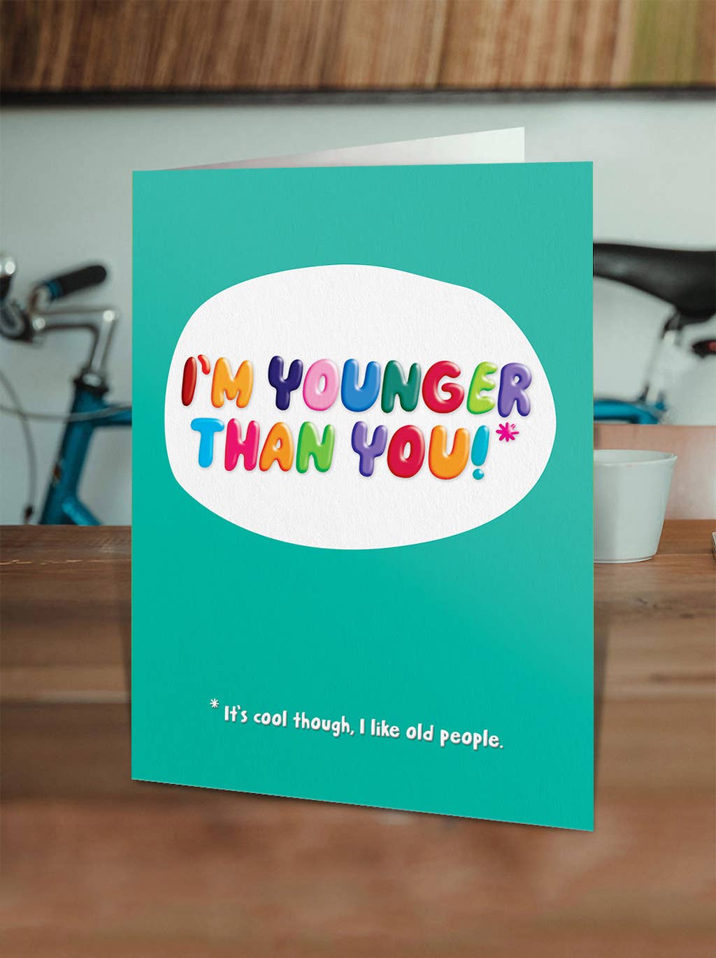 Teal card with colorful text 'I'm younger than you!' on a wooden surface.