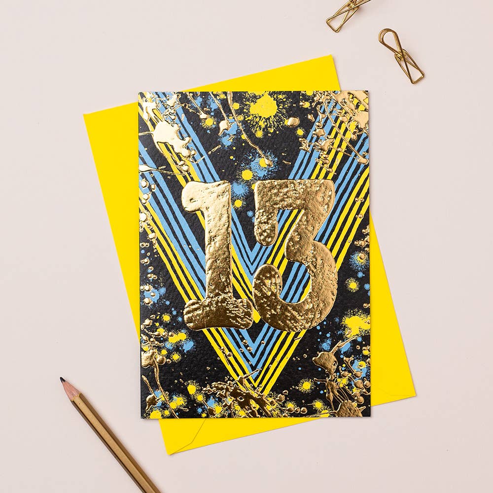 A colorful birthday card with gold foiling and the number 13 in the center, placed on a yellow envelope, with a pencil and paper clips on the side.