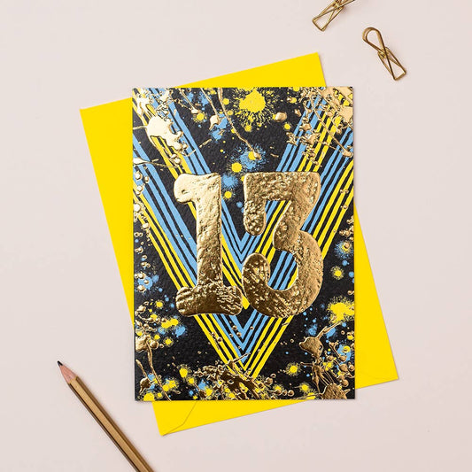 A colorful birthday card with gold foiling and the number 13 in the center, placed on a yellow envelope, with a pencil and paper clips on the side.