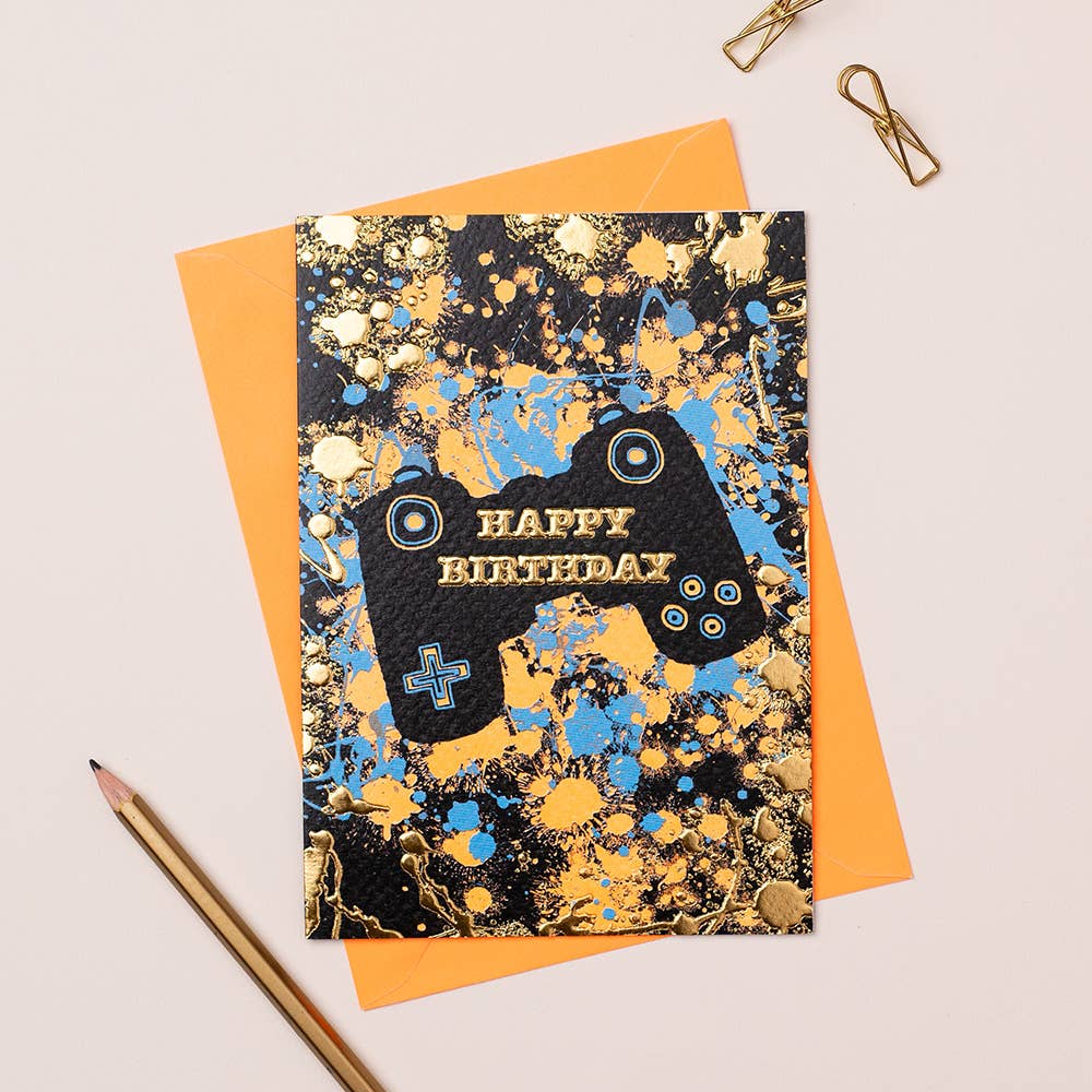 A greeting card with a gaming-themed design, featuring a gold and blue splatter pattern and the words 'Happy Birthday' on the front, accompanied by an orange envelope and a pencil on a white surface.