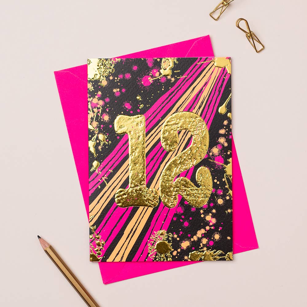 A vibrant birthday card with gold foiling and the number 12 in a bold, decorative font, accompanied by a pink envelope and a pair of gold-colored pencils on a white background.