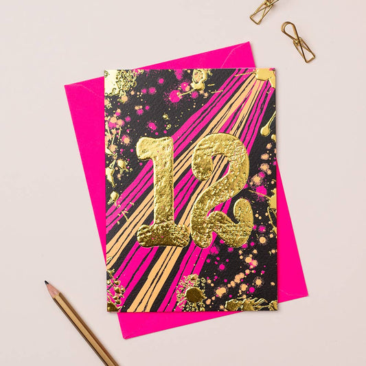 A vibrant birthday card with gold foiling and the number 12 in a bold, decorative font, accompanied by a pink envelope and a pair of gold-colored pencils on a white background.