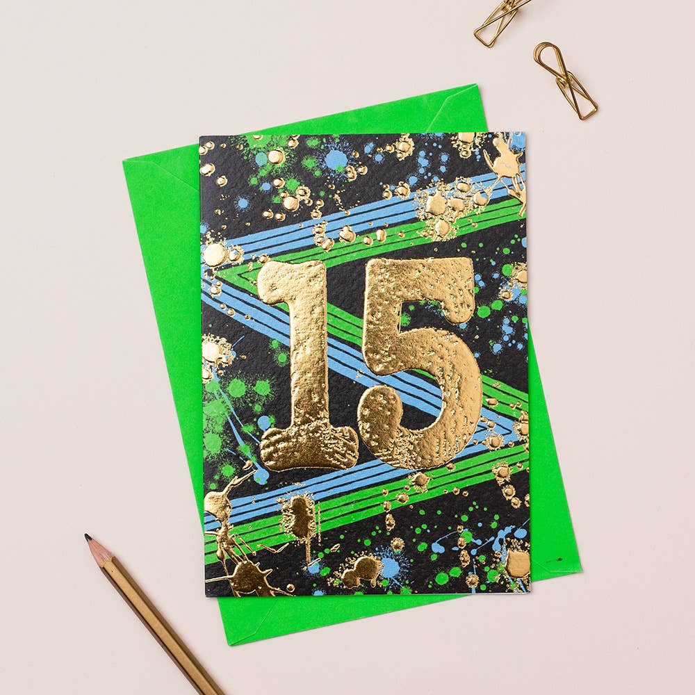 A greeting card with a colorful design, featuring gold foiling and the number 15, presented alongside a green envelope and a pencil.