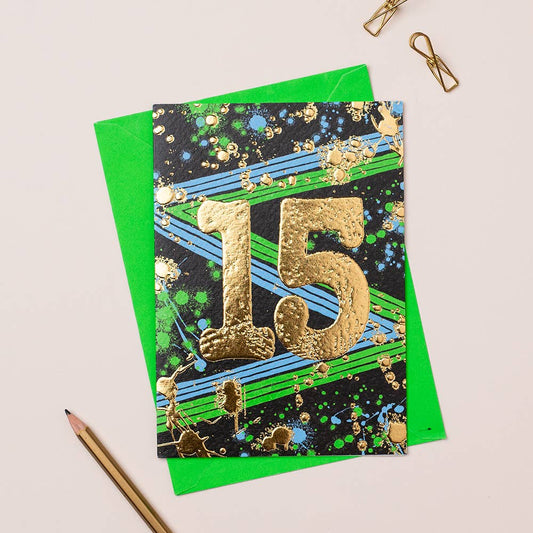 A greeting card with a colorful design, featuring gold foiling and the number 15, presented alongside a green envelope and a pencil.