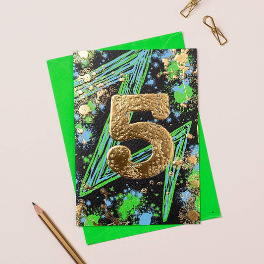 A vibrant birthday card with a gold number 5 on a black background, positioned on a table with a pencil and a clipstone.