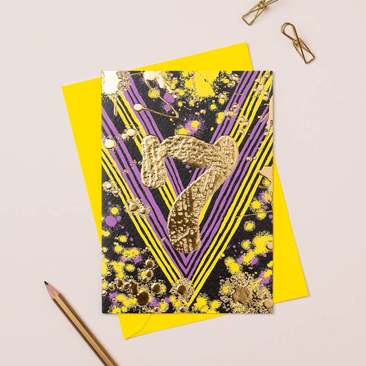A colorful birthday card with gold foiling and a geometric design, placed on a yellow envelope, accompanied by a pencil and a clip on a white surface.