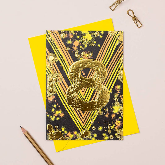 A colorful birthday card with gold foiling and the number 8, placed on a yellow envelope, displayed with a pencil and paper.
