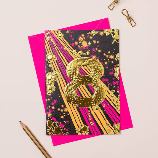 A vibrant birthday card with gold foiling, designed for an 8-year-old girl, featuring a black background with pink and gold patterns, and a large gold '8' in the center. The card is presented with a pink envelope and a pair of gold-colored pins on a white surface.