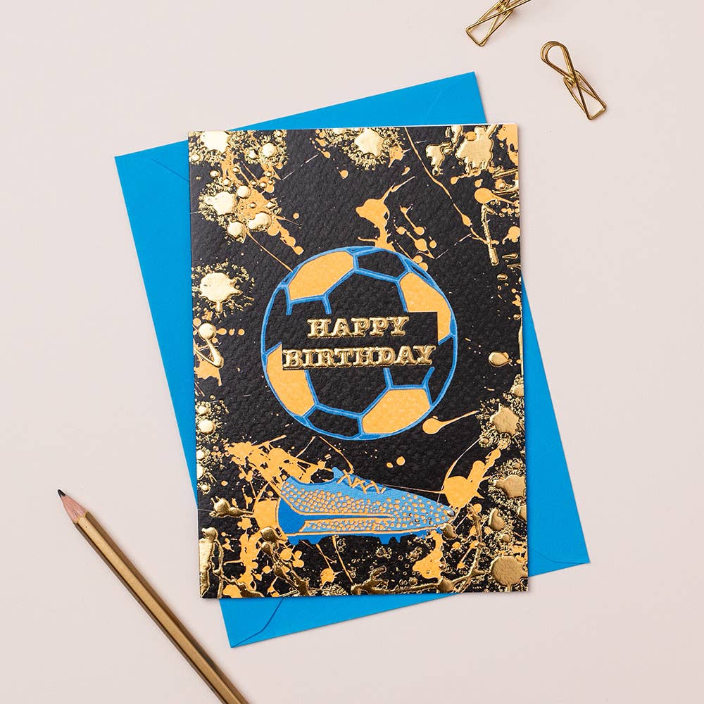 A birthday card with a football splatter design, featuring gold foiling and the text 'Happy Birthday' on a blue background, accompanied by a blue envelope and a pair of gold-colored pencils.