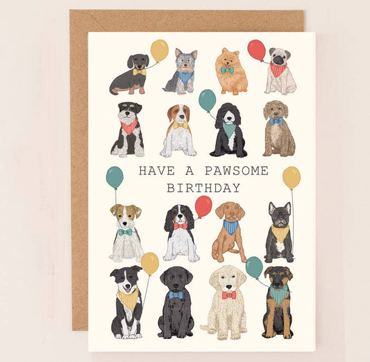 A birthday card with various dog illustrations, each wearing a bow tie and holding a balloon, with the text 'HAVE A PAWSOME BIRTHDAY' at the center. The card is accompanied by a brown kraft envelope.