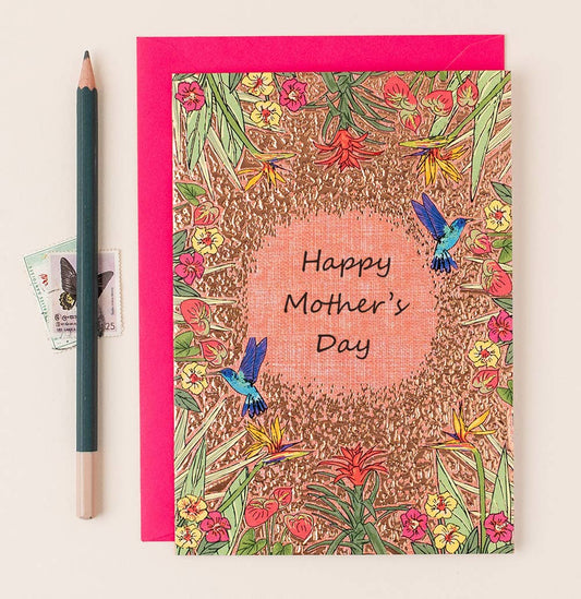 A Mother's Day card with a gold foil border and tropical flower design, featuring hummingbirds and the text 'Happy Mother's Day'. Accompanied by a red envelope and a pencil, all placed on a white surface.