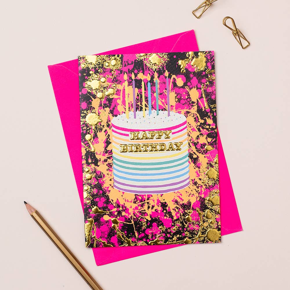 A vibrant birthday card with a gold foiled cake and 'Happy Birthday' text on a pink background, accompanied by a blue envelope and a pencil.