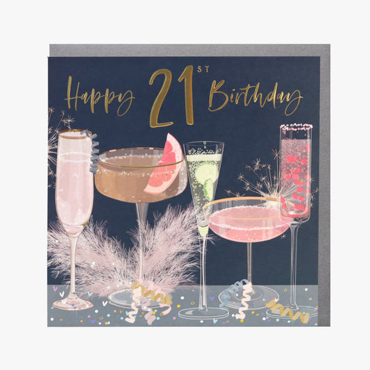 A greeting card with a 21st birthday theme, featuring illustrations of three cocktails and a 'Happy 21st Birthday' message, embellished with gold foiling and set against a dark background. The card is presented with a silver envelope.
