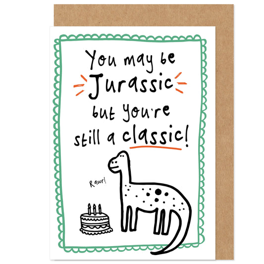 A birthday card with a dinosaur illustration, featuring text that reads 'You may be Jurassic but you're still a classic!' with a brown kraft envelope.