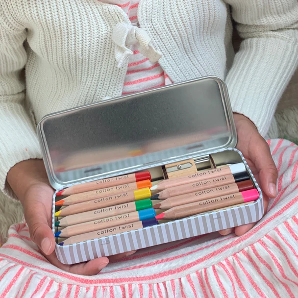 A little girl in a pink striped dress and white cardigan holding an open tin of Colouring pencils.