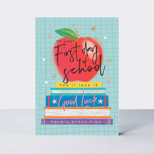 A greeting card with an illustration of an apple and books, titled 'First Day of School' with a wish for good luck.