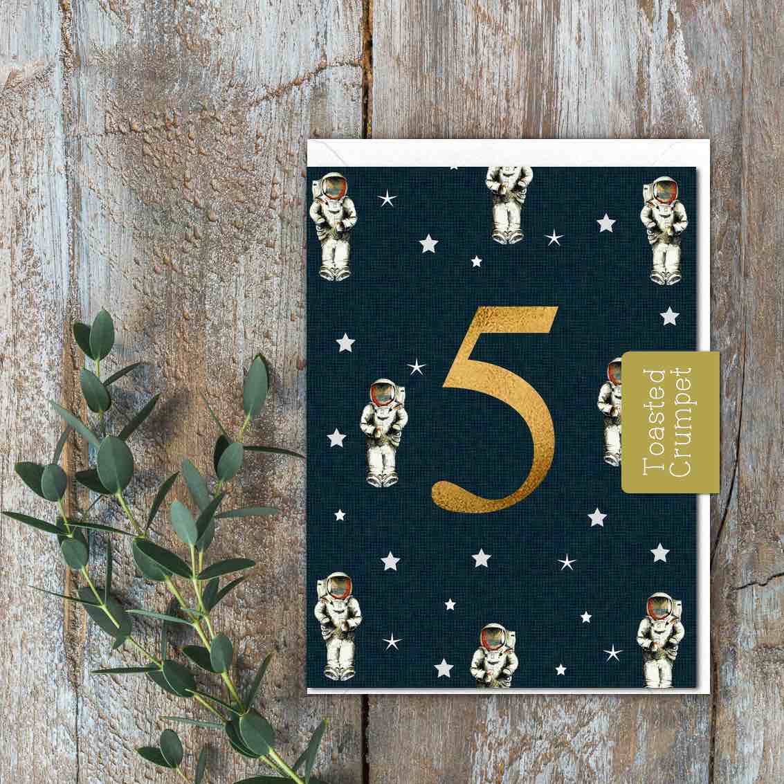 A small black card with stars and astronauts on with a large gold 5 in the middle of the card.