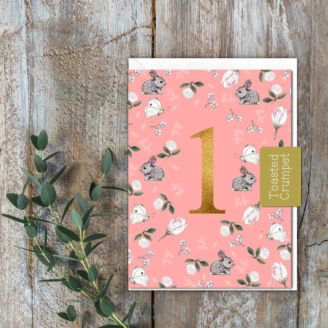 A small pink card with bunnies, white flowers and butterflies and a large gold one in the centre.