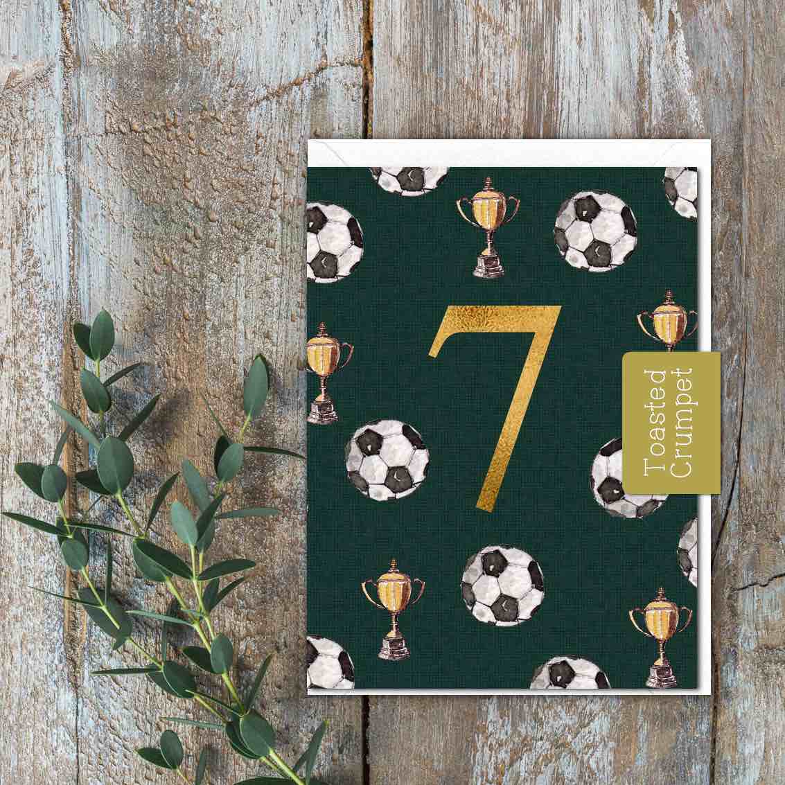 A mini dark green card with a gold age 7 on it. Footballs and trophies on the card.