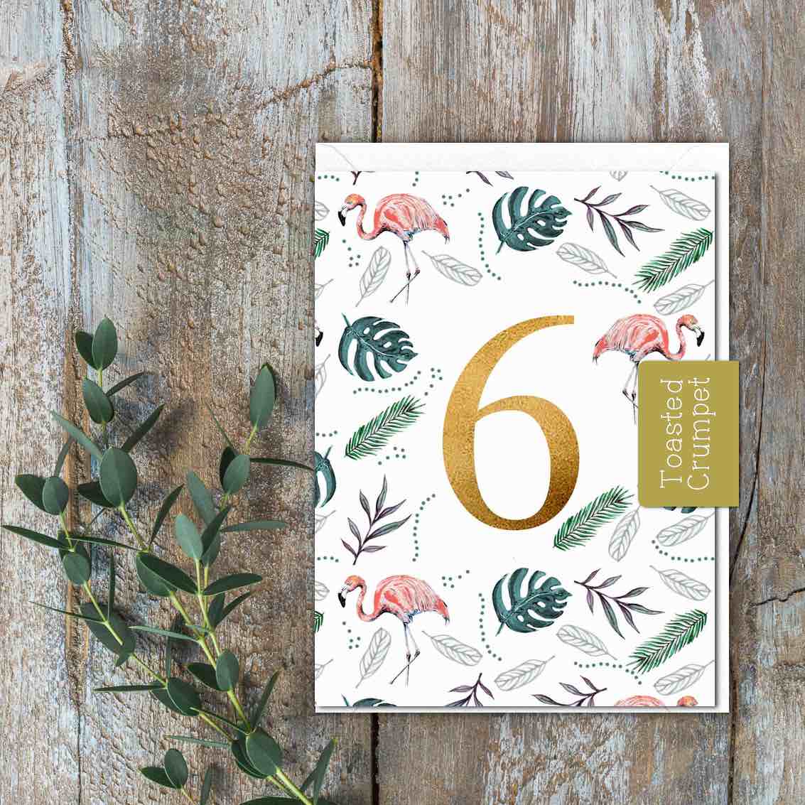 A small white card with flamingos and leaves with a large gold 6 in the middle of the card.