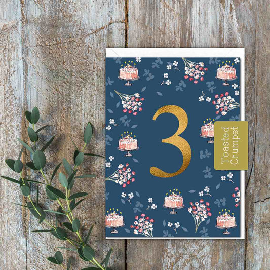 A small blue card with pink flowers and cakes and a gold 3 in the middle.