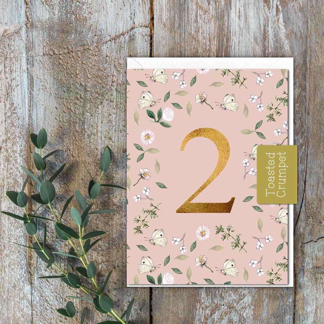 A small pink card with white and green flowers and butterflies and a large gold 2 in the middle/